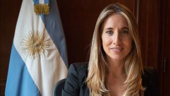 headshot of a women against a dark brown wall, with the Argentinian flag in the background. She has long, dark blonde hair and she is wearing a black suit jacket. She is smiling. 
