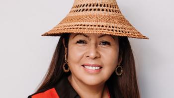 Headshot of a smiling women against an offwhite background. She is smiling and has long brown hair. She is wearing a light brown hat and small circle earrings. 
